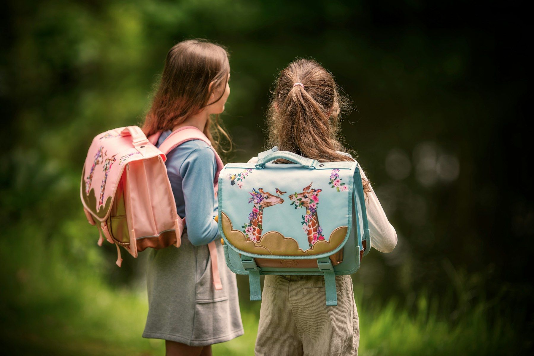 Zwei Mädchen mit Schulranzen, von hinten, auf dem Weg durchs Grüne.